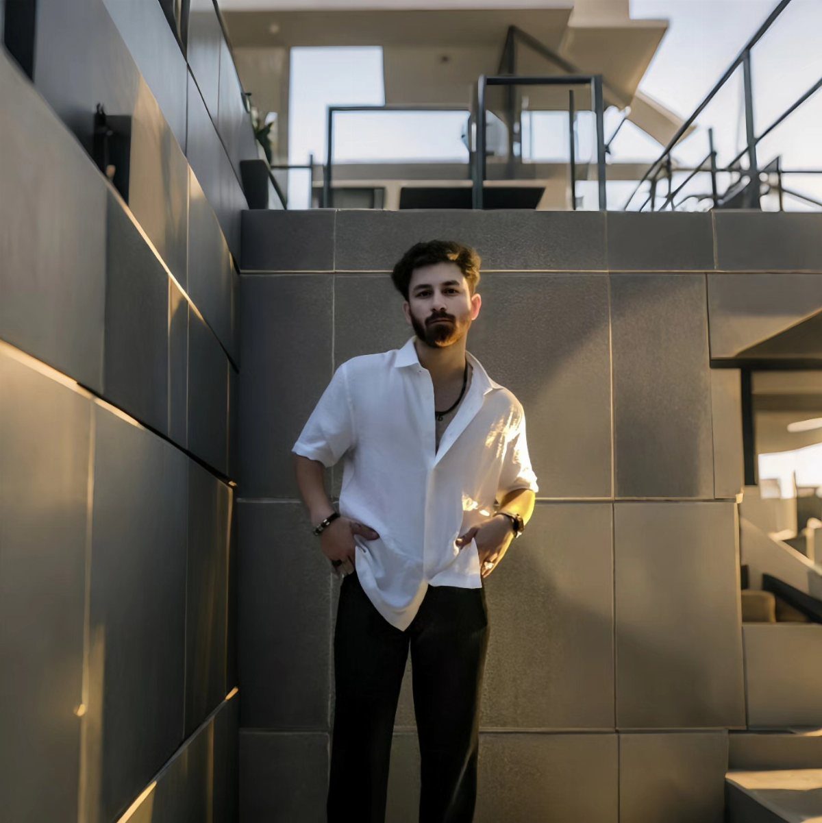 Man in a white shirt and black trousers posing against a modern grey tiled wall.