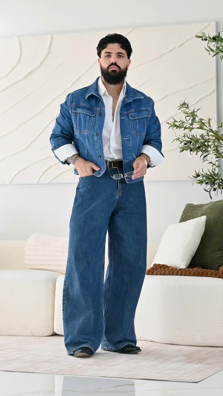 A bearded man posing in a denim jacket, white shirt, and wide-leg blue jeans.
