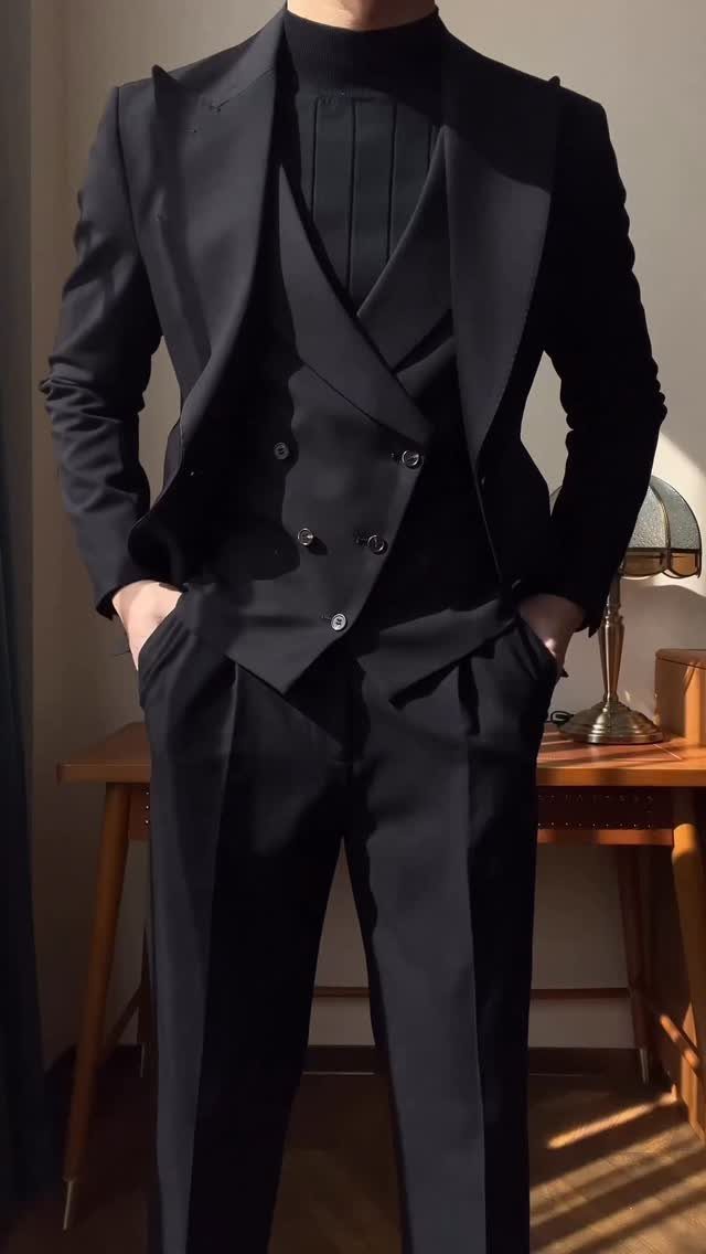 A man dressed in a sharp, black three-piece suit and turtleneck standing indoors.