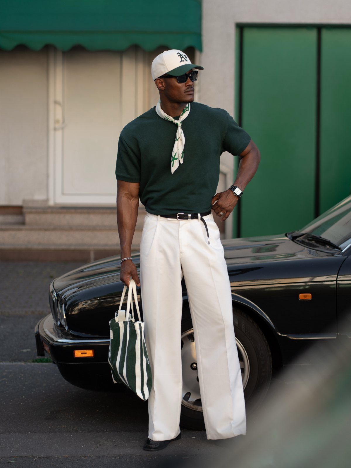 Man in green t-shirt and white trousers posing next to a black car.