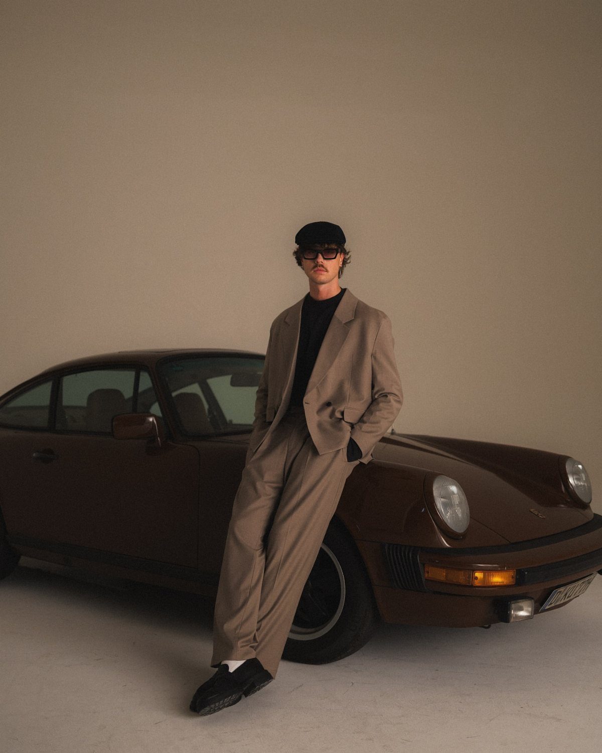 A man in a tan oversized suit and black cap leans against a vintage brown sports car.