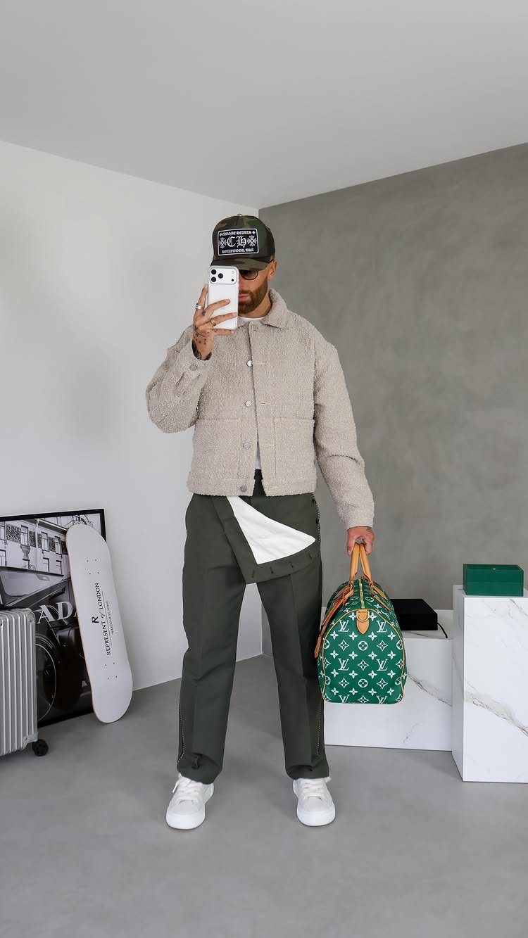 Man in beige jacket, olive trousers, and trucker hat holding a green monogrammed bag.