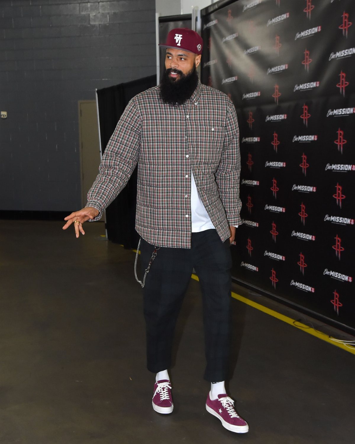 A bearded man in a plaid shirt, dark pants, and maroon sneakers walking through a hallway.
