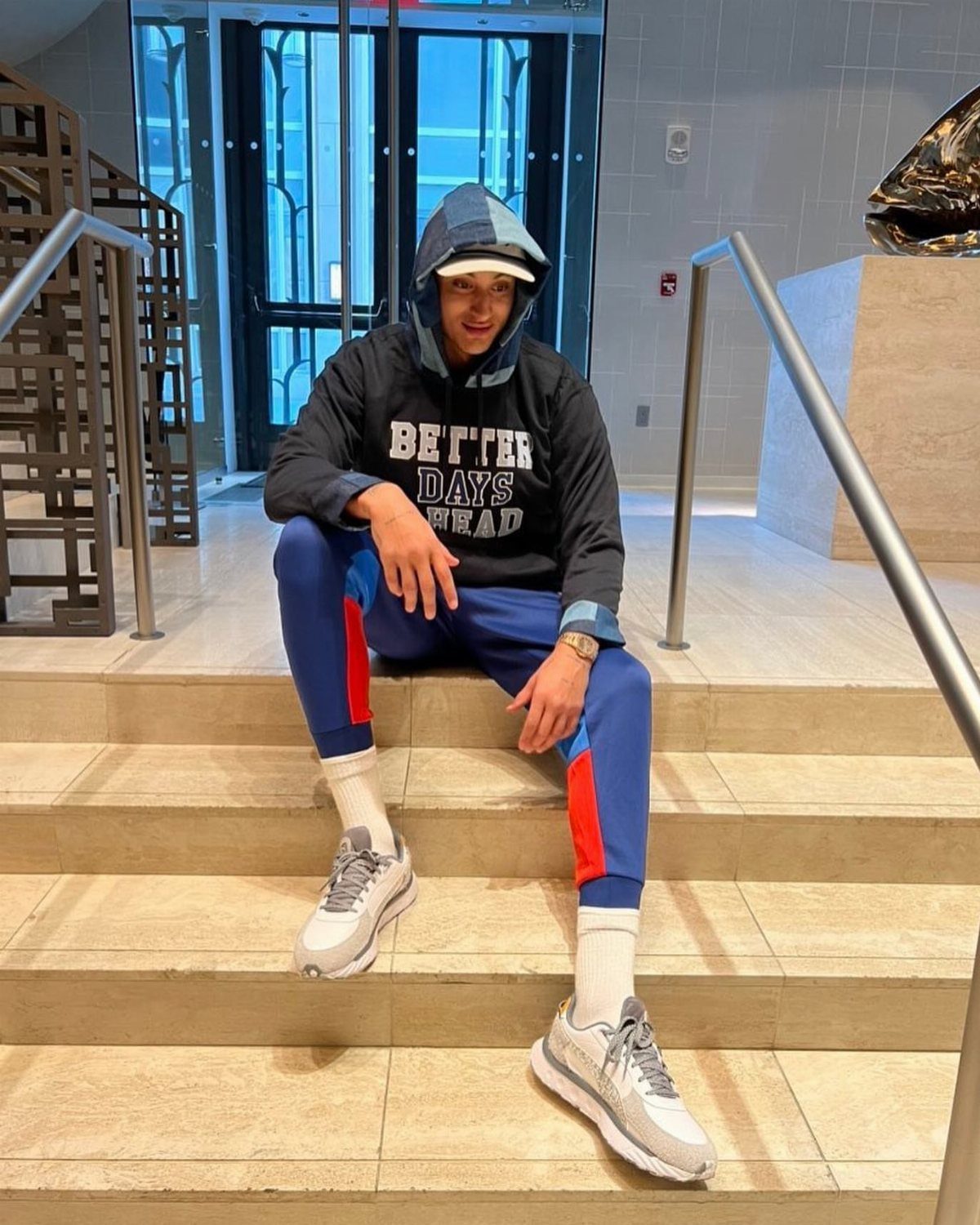 Man in black hoodie and blue athletic pants sitting on stairs wearing grey sneakers.