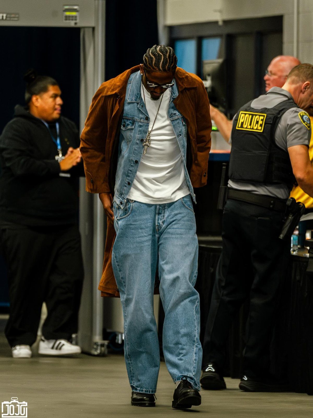 A man in a brown suede coat and baggy denim outfit walks through a hallway.