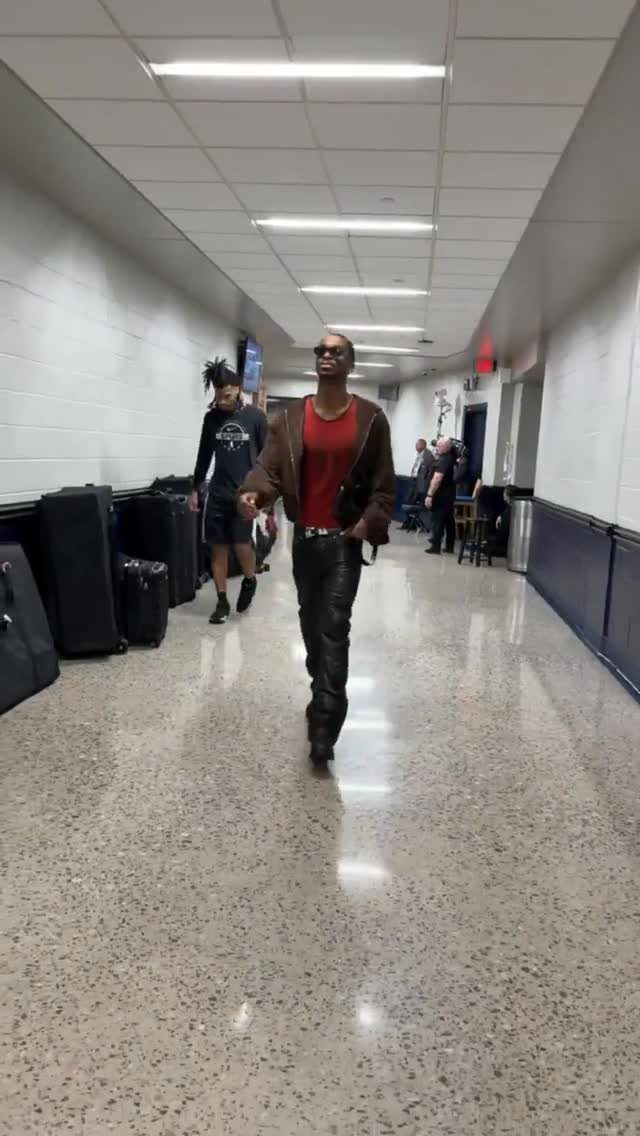 A man in leather pants and a brown jacket walks through a long arena hallway.