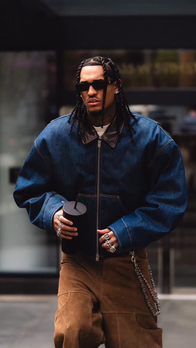 A man in an oversized denim jacket and brown trousers walking while wearing sunglasses.