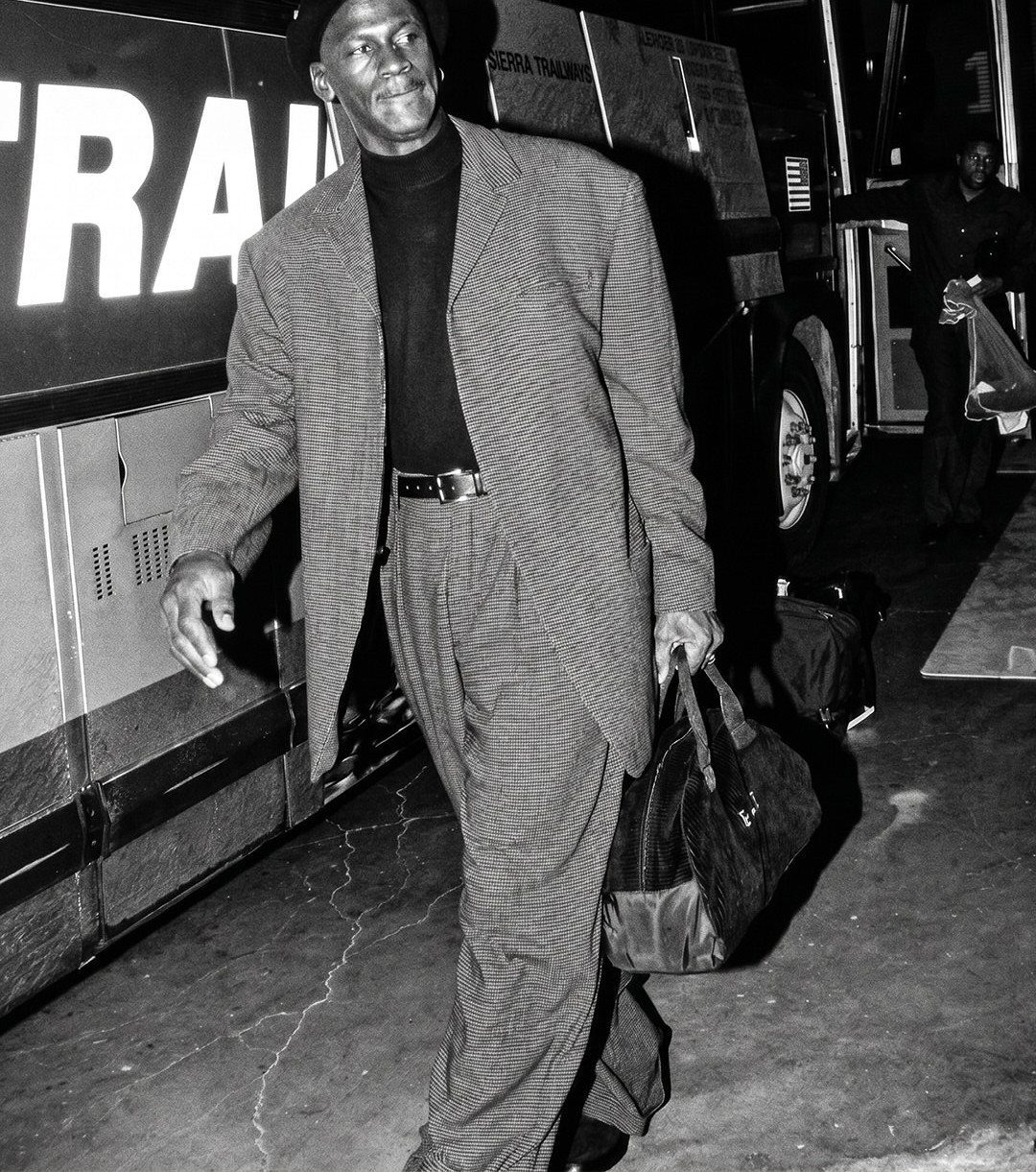 A man in a patterned suit and beret walks past a large vehicle at night.