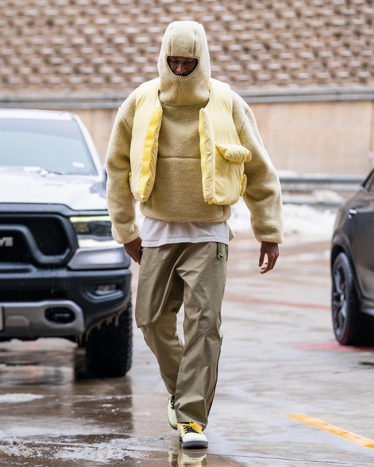 Man walking in a cream fuzzy hooded sweater and yellow utility vest with tan trousers.