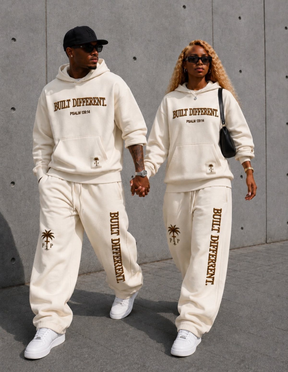 Man and woman in matching cream streetwear walking hand-in-hand against a concrete wall.