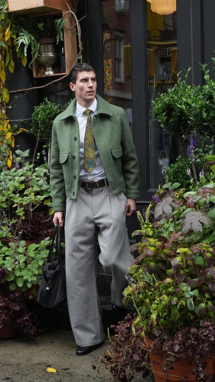 A man wearing a green jacket, patterned tie, and wide-leg grey trousers standing among potted plants.