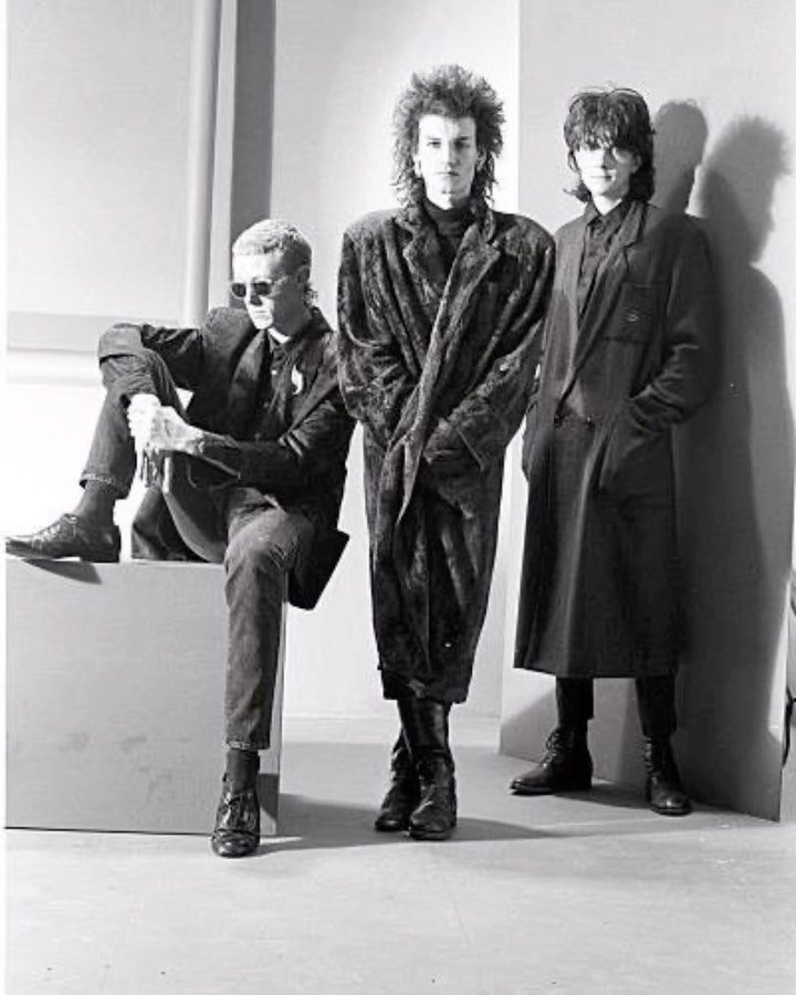 Three men posing in 1980s new wave style clothing and long coats.
