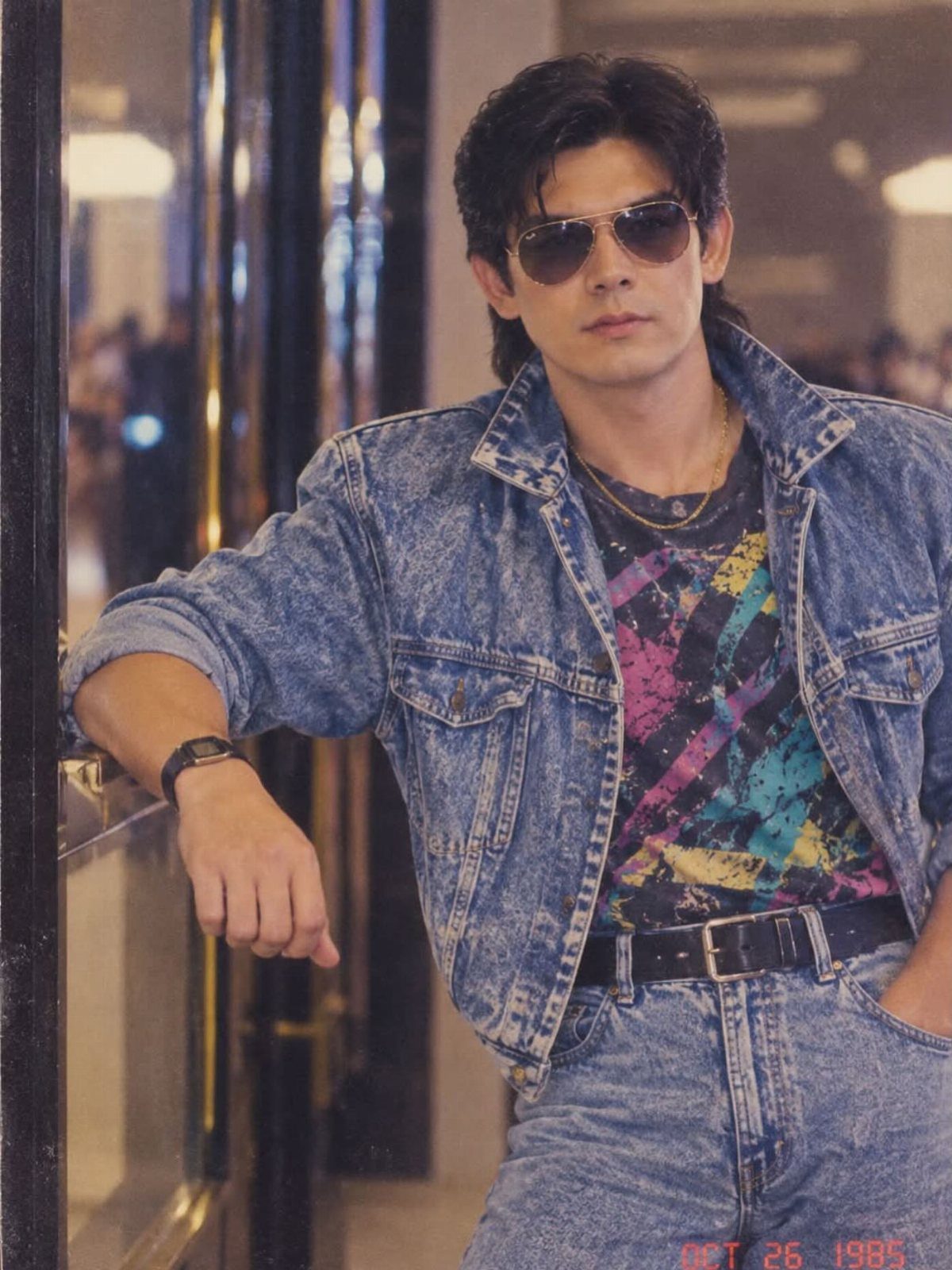 Man in sunglasses wearing an oversized denim jacket and colorful graphic t-shirt leaning against a door.