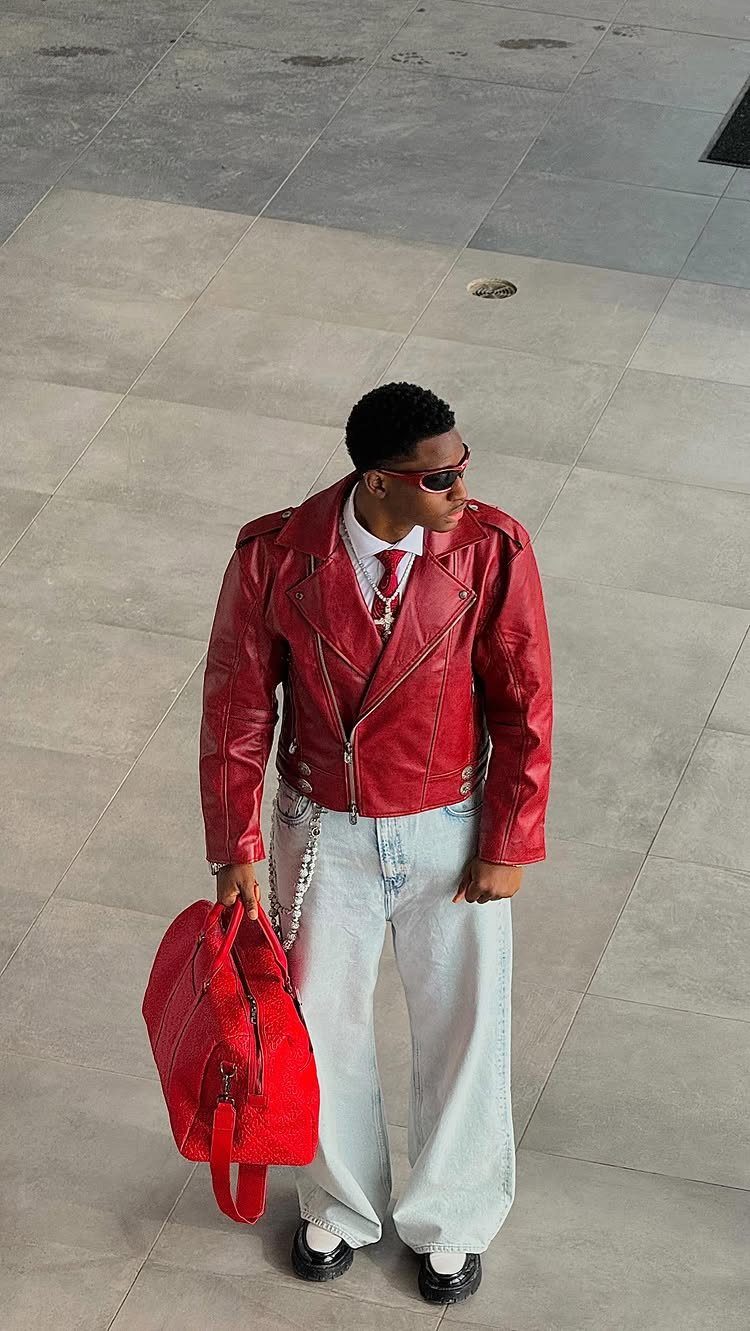 A person in a red leather jacket, red tie, and wide-leg jeans standing on pavement.