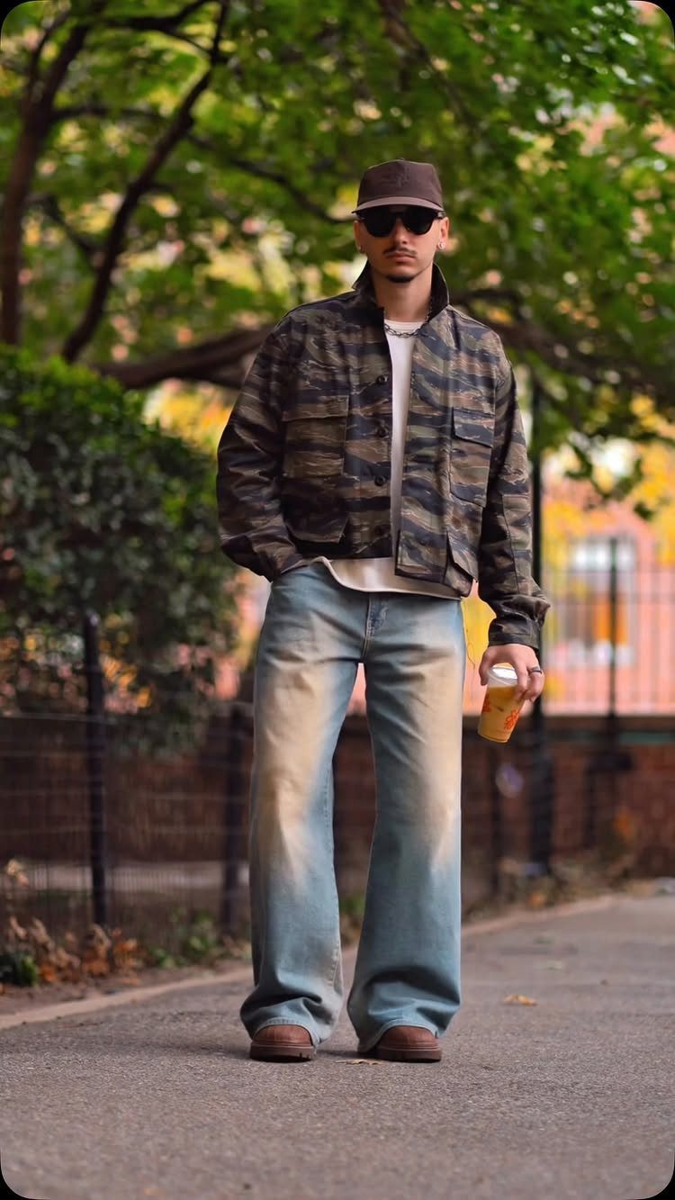 A man in a camouflage jacket and wide-leg jeans standing on a city sidewalk near trees.