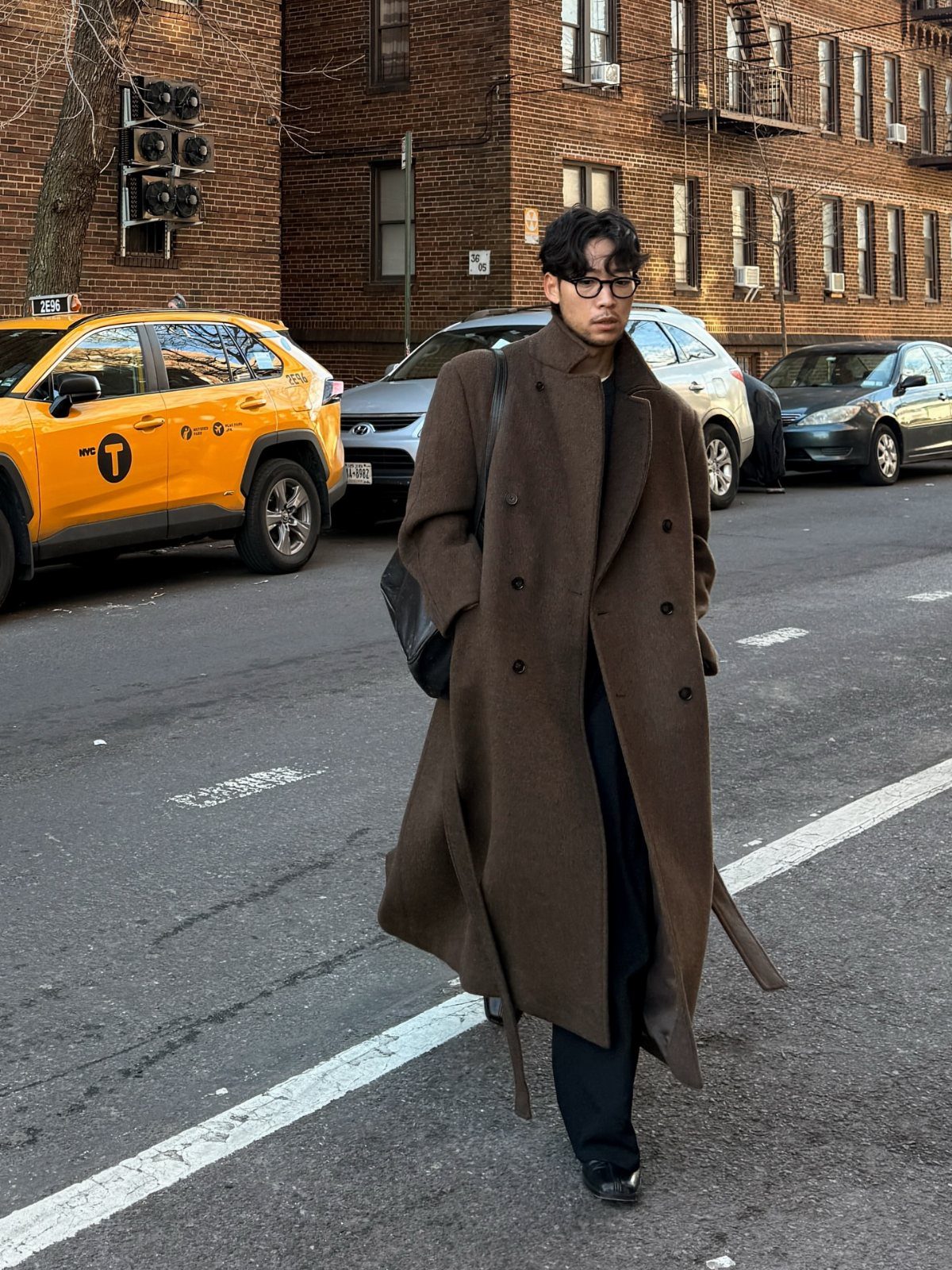 Man walking down a city street wearing a long brown wool overcoat and dark trousers.