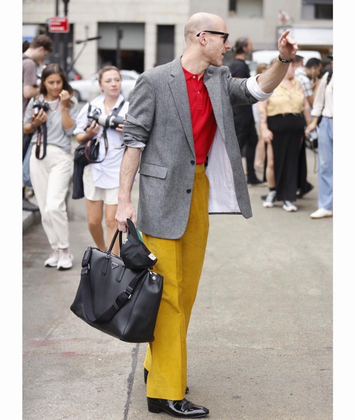 A man walking in a city wearing a grey blazer, red shirt, and yellow trousers while carrying a black bag.