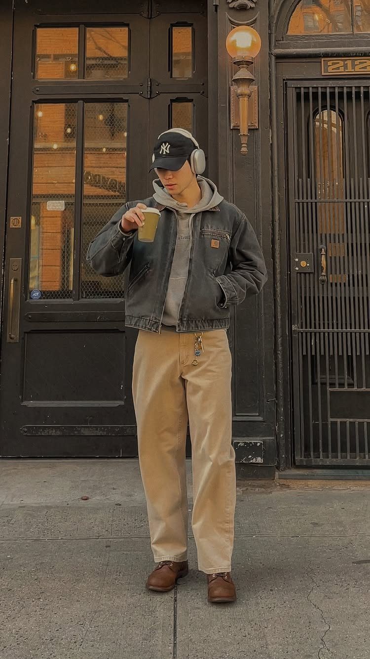 A man in a grey jacket, tan pants, and a baseball cap stands on a city street.