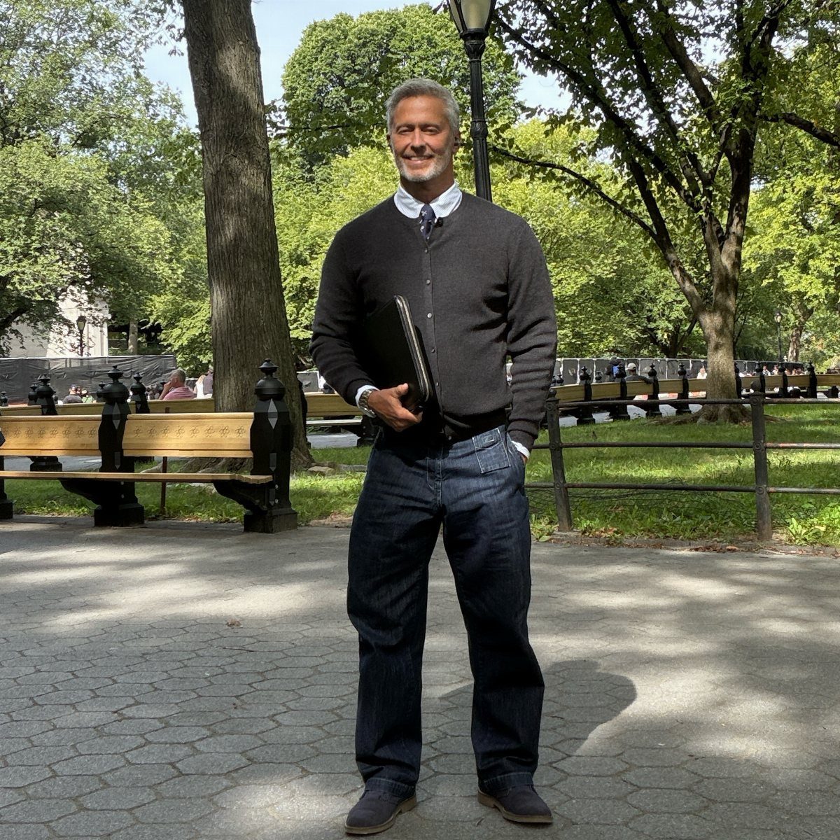 A smiling man in a black cardigan and dark denim jeans standing in a sunny park.