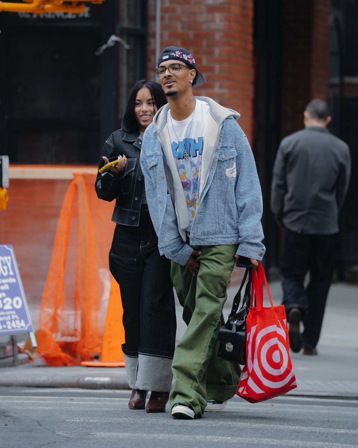 Man in denim jacket and green trousers walking through a city street with a woman.