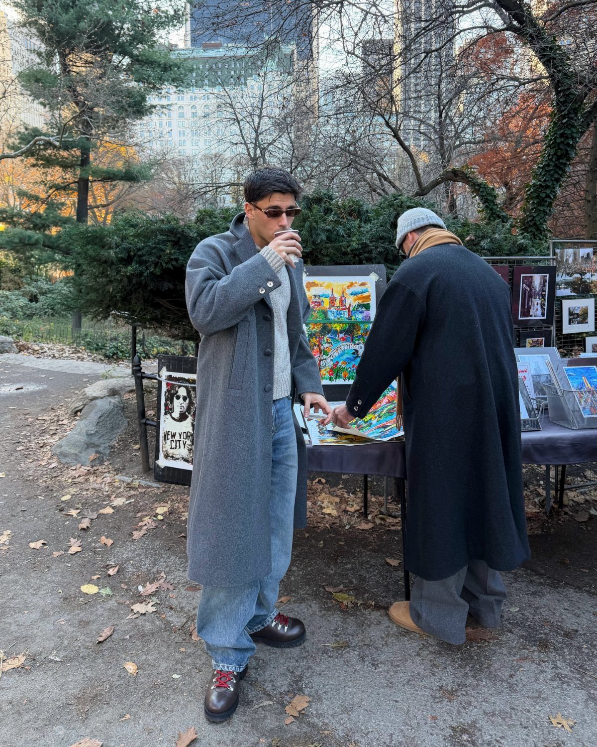 Man wearing a long grey wool coat, sweater, and blue jeans in a park setting.