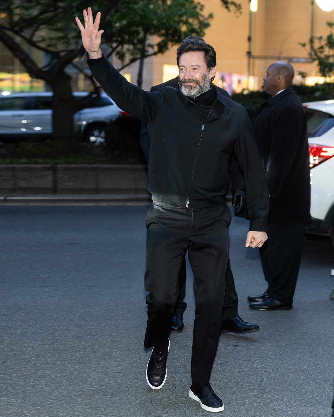 Man in an all-black jacket and trousers waving while walking outdoors at night.