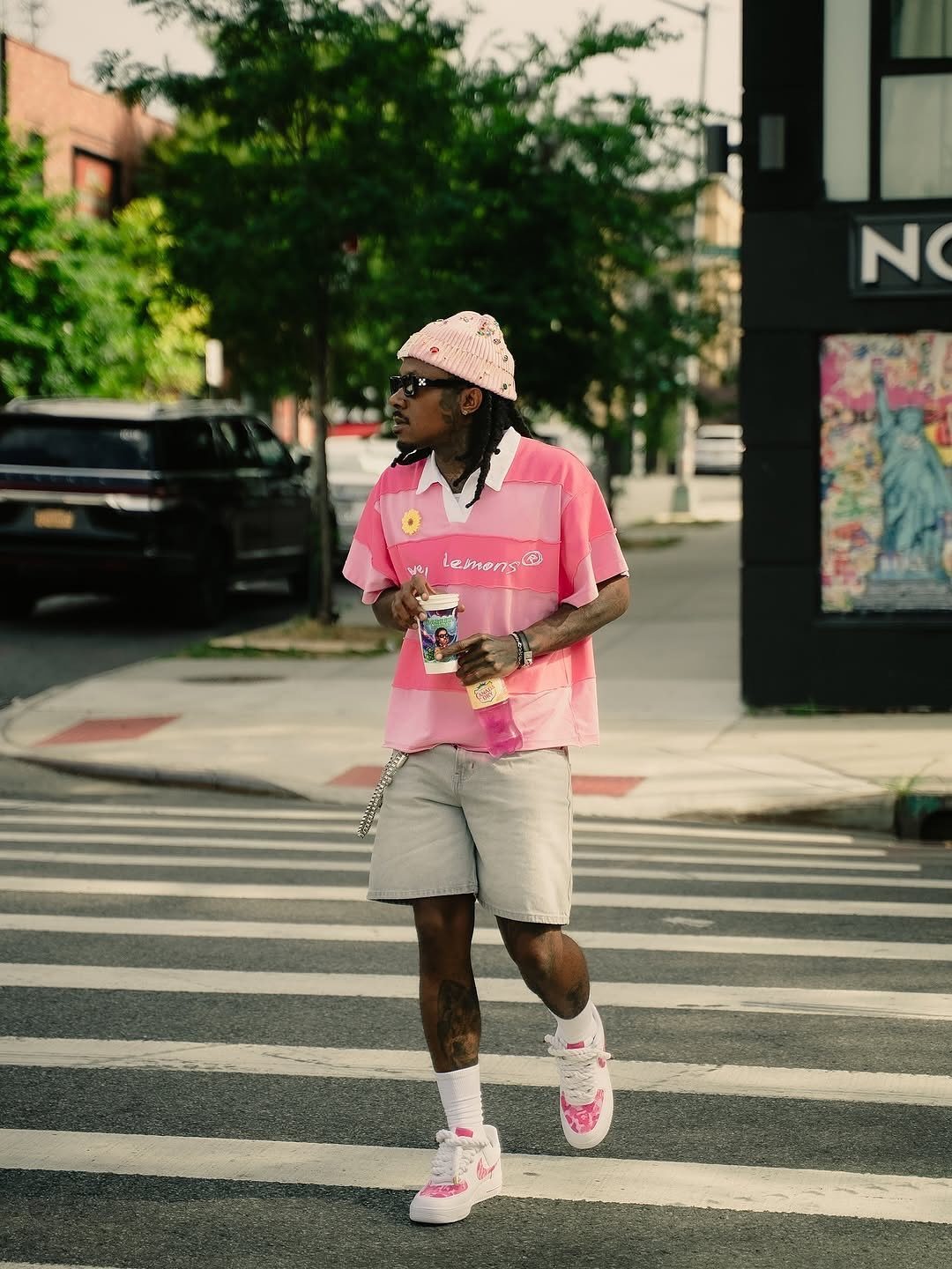 A man in a pink striped polo and grey shorts walks across a city street crosswalk.