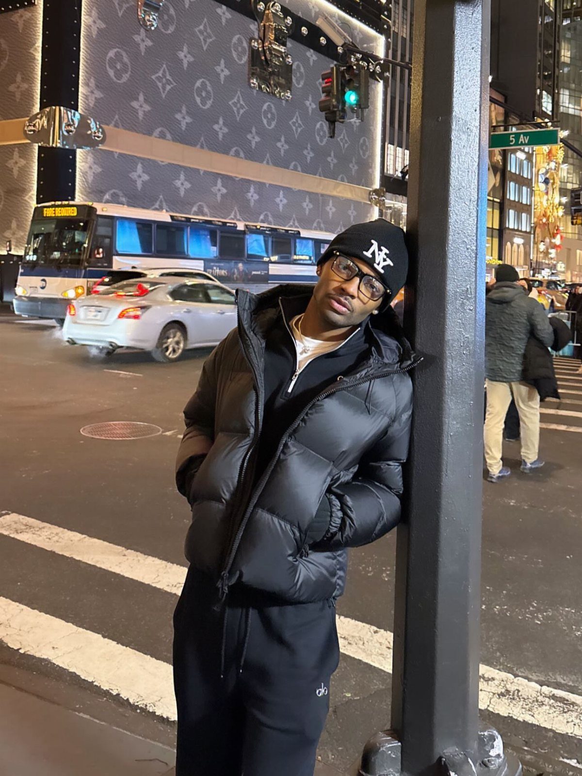 Man in a black puffer jacket and beanie leaning against a pole on a city street at night.