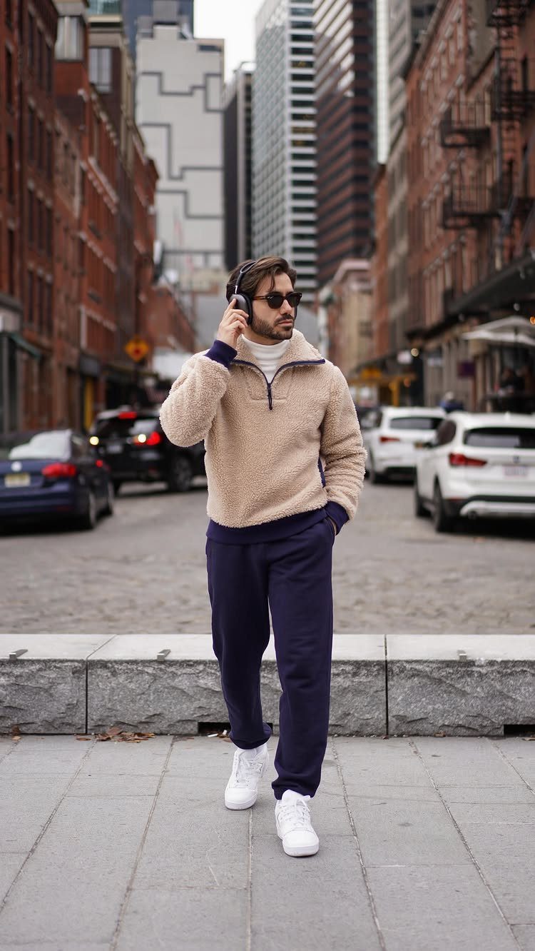 Man in beige fleece and navy sweatpants walking on a city street wearing headphones and sunglasses.
