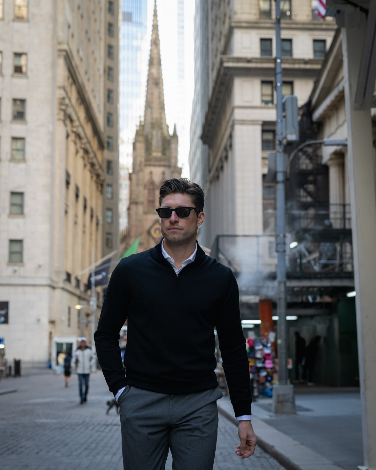 Man in black sweater and grey trousers walking through a city street with tall buildings.