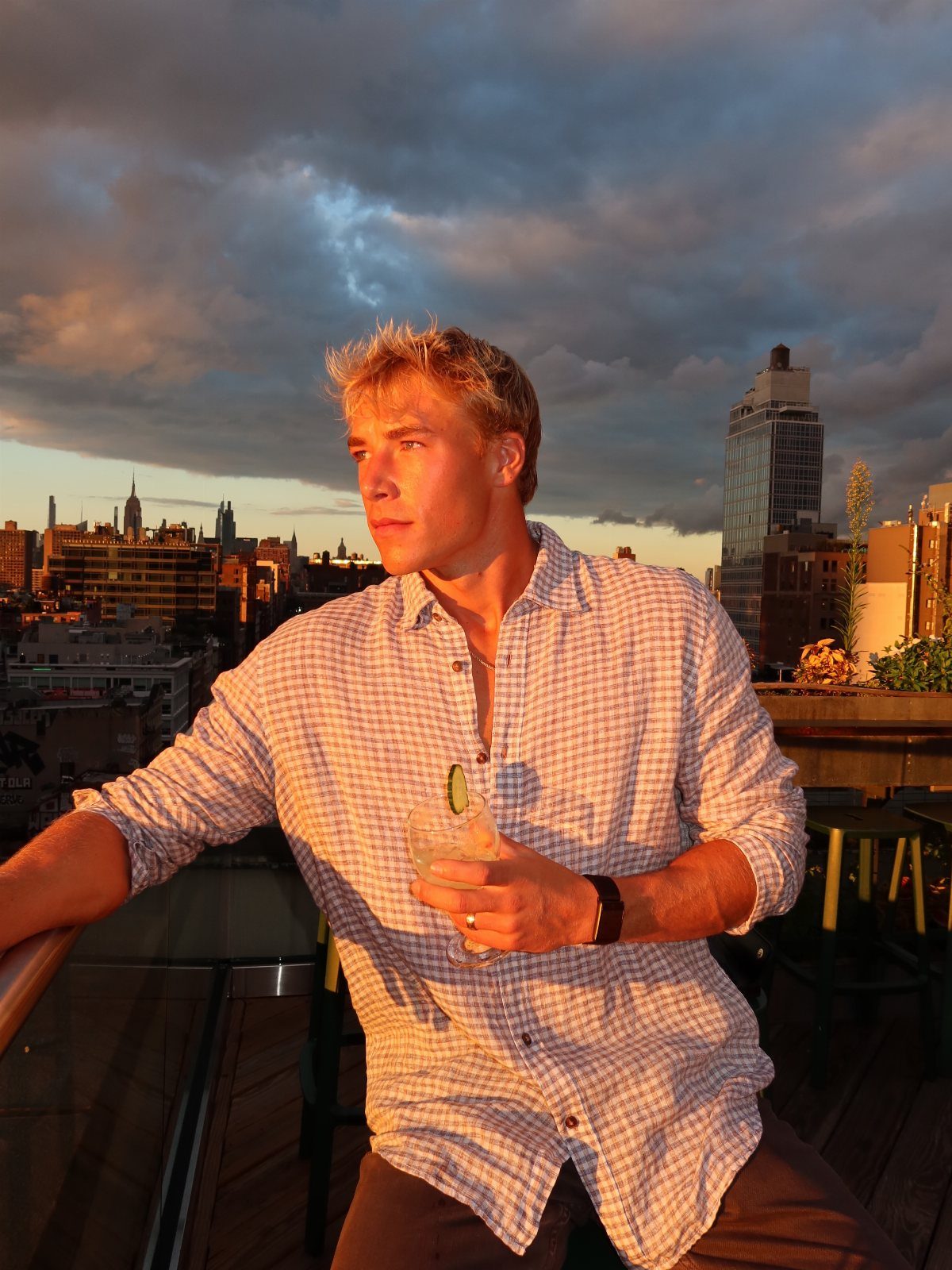 Man in a checkered shirt holding a cocktail on a city rooftop at sunset.
