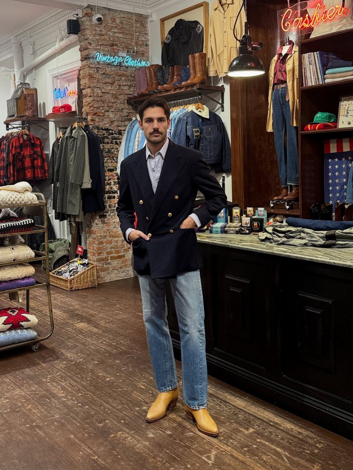 Man posing in a vintage shop wearing a navy blazer, light jeans, and tan boots.