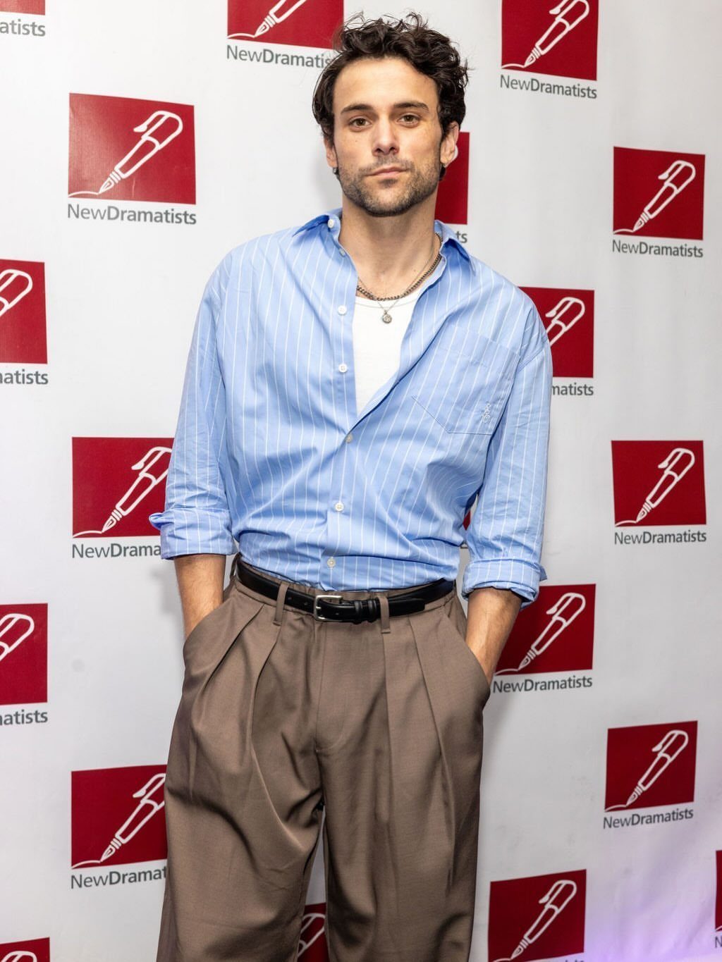 Man in a blue striped shirt and brown pleated trousers posing against a branded backdrop.