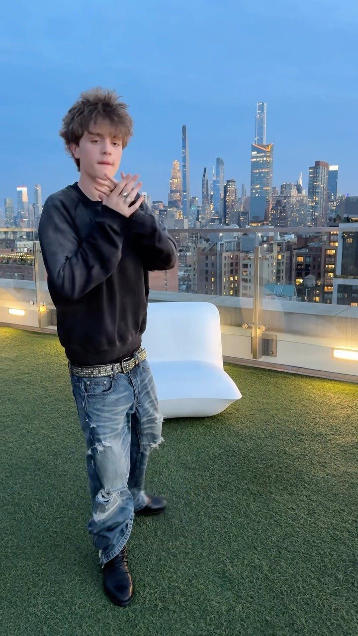 Young man in black sweatshirt and distressed jeans posing on a rooftop overlooking a city skyline.