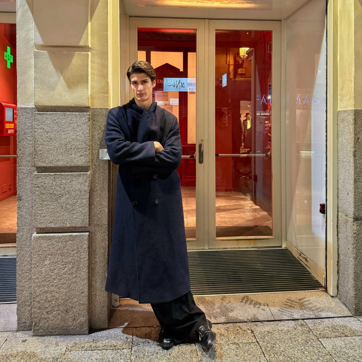 Man in a long navy overcoat and black trousers posing against a stone pillar.