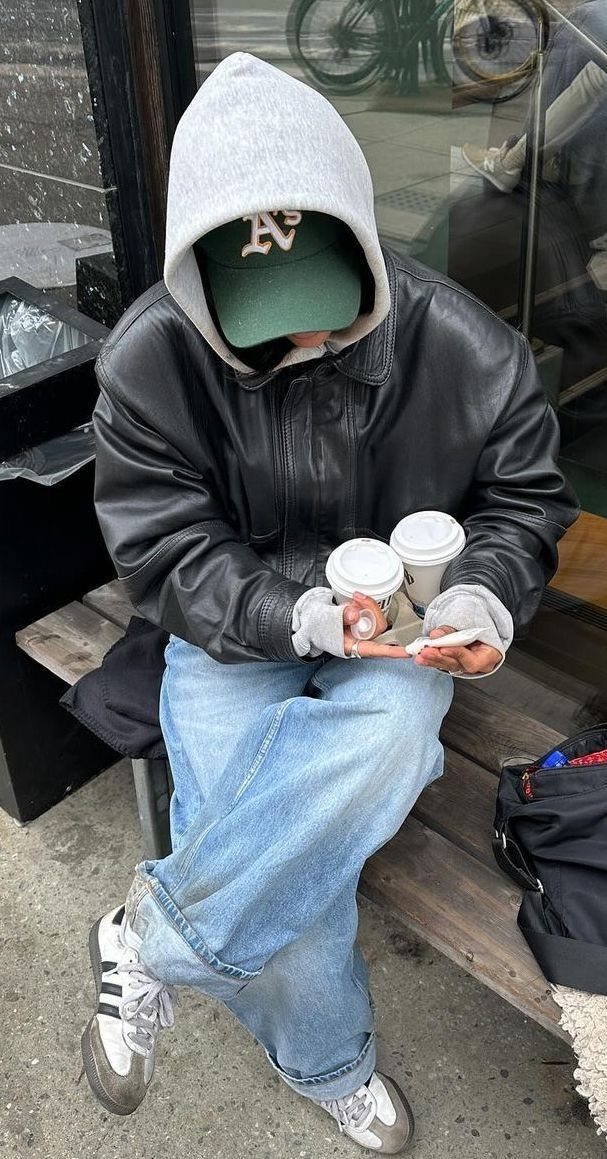 A person wearing a leather jacket, grey hoodie, green cap, and baggy jeans sitting on a bench.