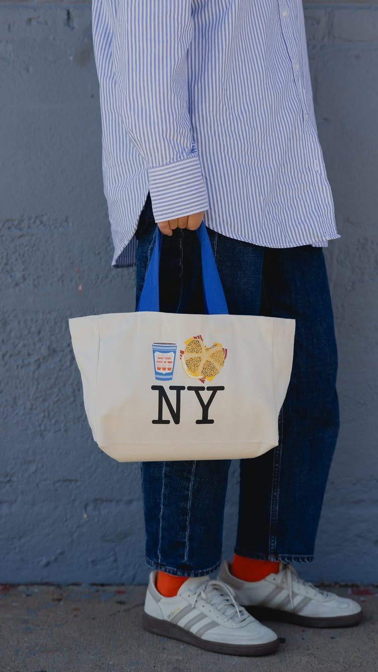 A person holding a white canvas tote bag with blue handles and NY printed on it.