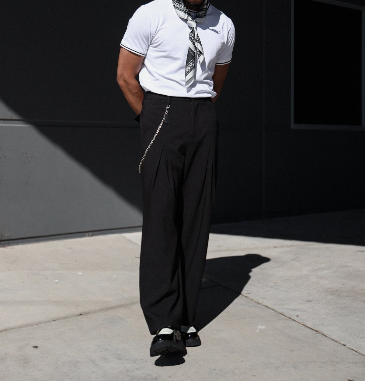 Person wearing a white t-shirt, black tailored trousers, and a patterned neck scarf standing outdoors.