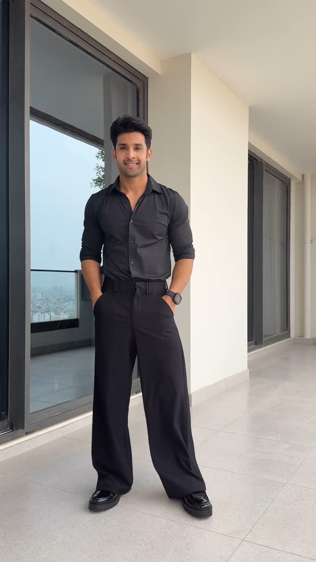 Man in a black button-down shirt and black wide-leg trousers standing on a balcony.
