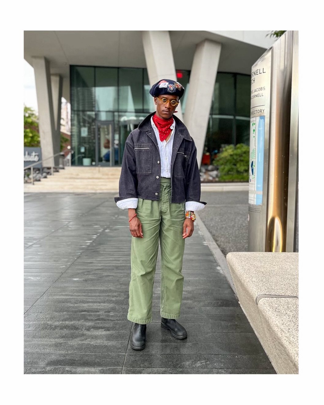 A man in a dark denim jacket, red bandana, and olive green trousers standing outdoors.