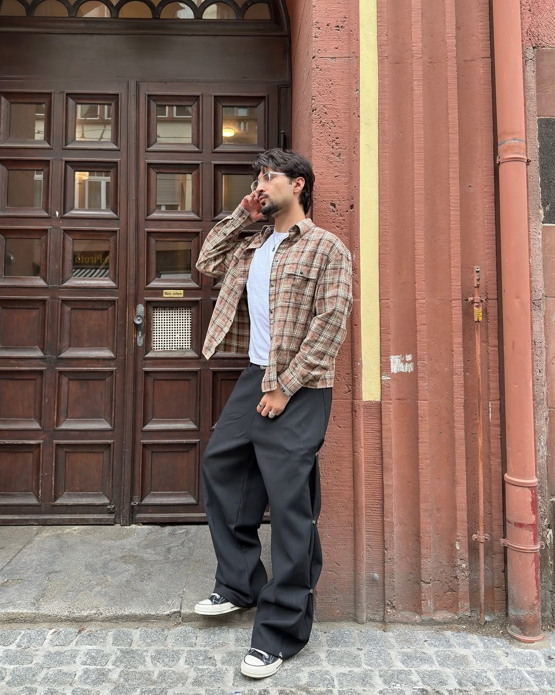 Man in a plaid shirt and wide-leg black pinstriped trousers standing by a wooden door.
