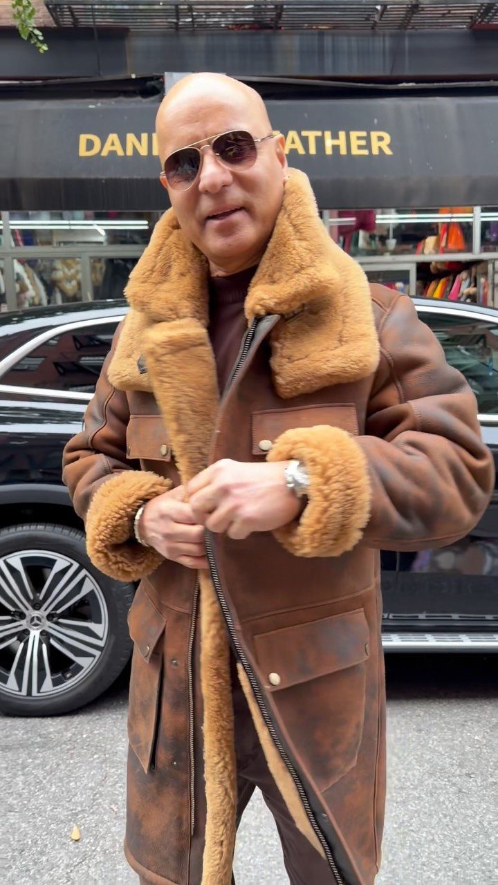 A man wearing a large brown shearling coat and sunglasses standing on a city street.