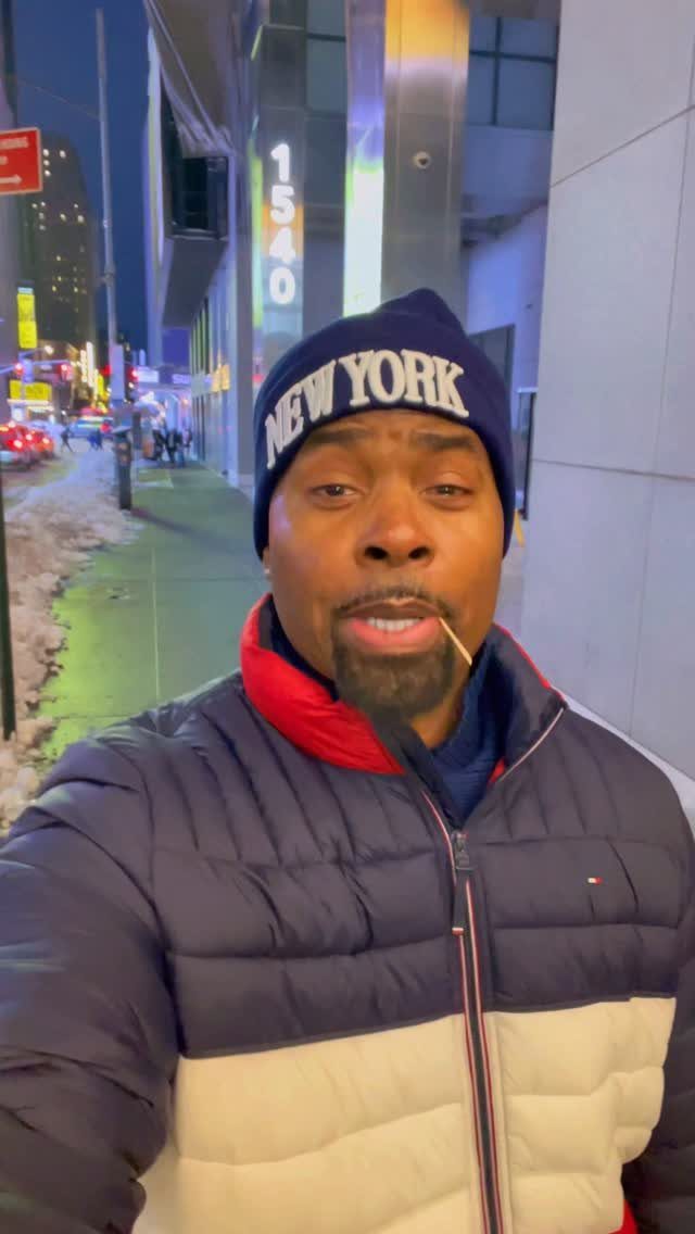 Man in a navy New York beanie and puffer jacket standing on a city sidewalk.