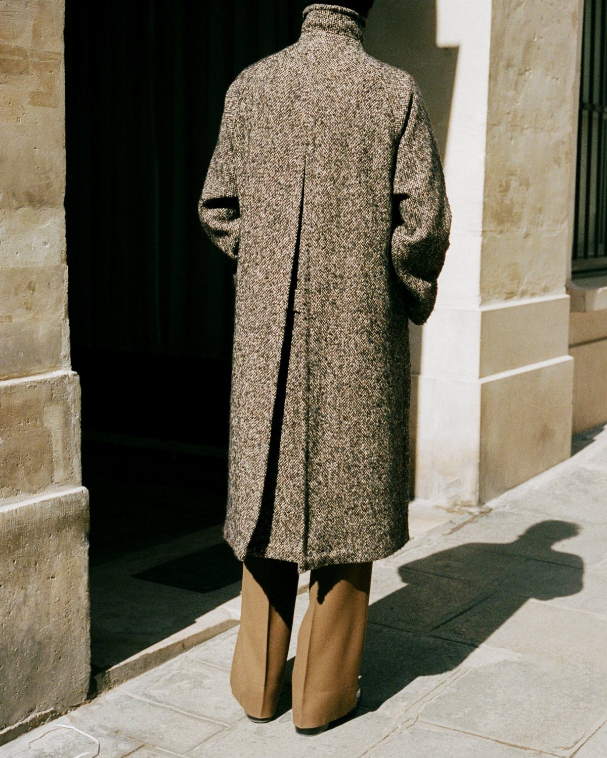 Person wearing a long brown tweed coat and tan wide-leg trousers standing outdoors.