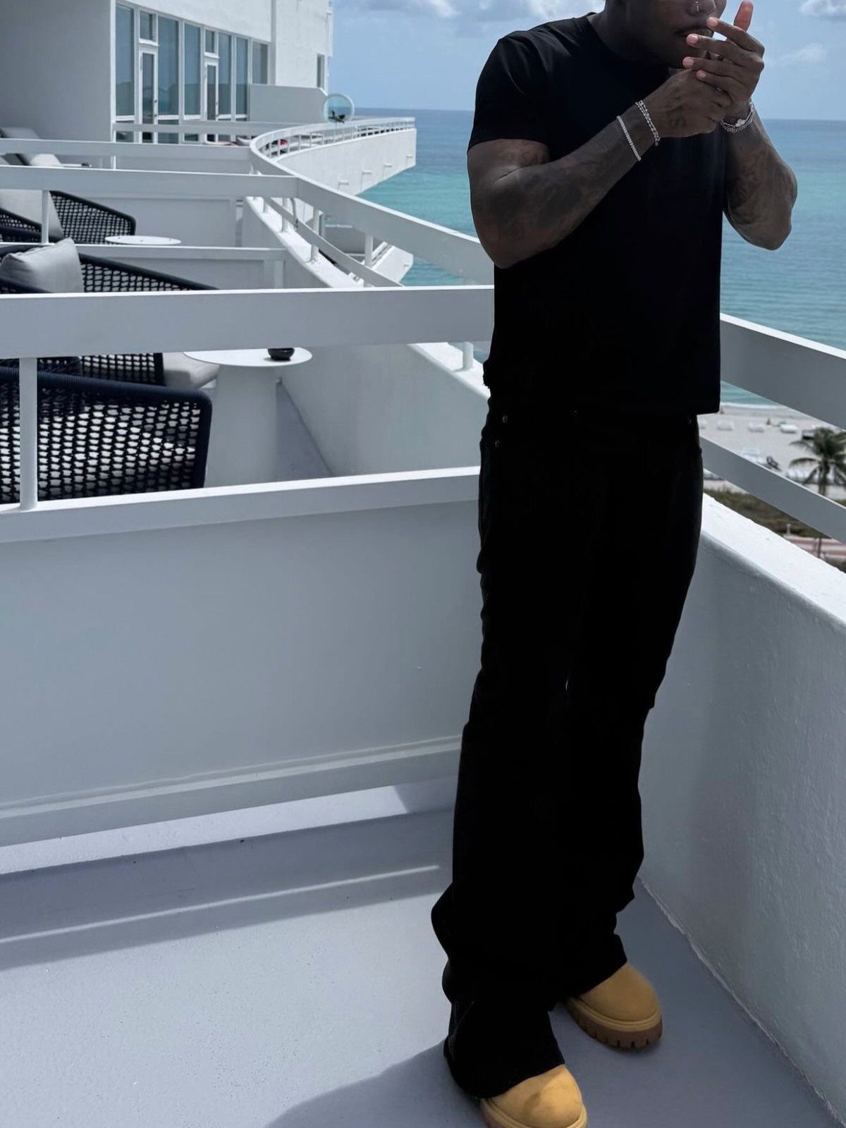 Man in an all-black outfit standing on a white balcony overlooking the ocean.