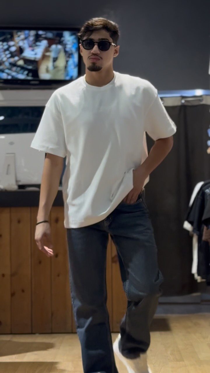 A man wearing a white t-shirt and dark blue jeans standing indoors.
