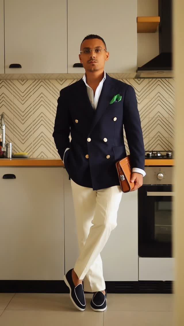 Man wearing a navy double-breasted blazer, white shirt, and cream trousers standing in a kitchen.