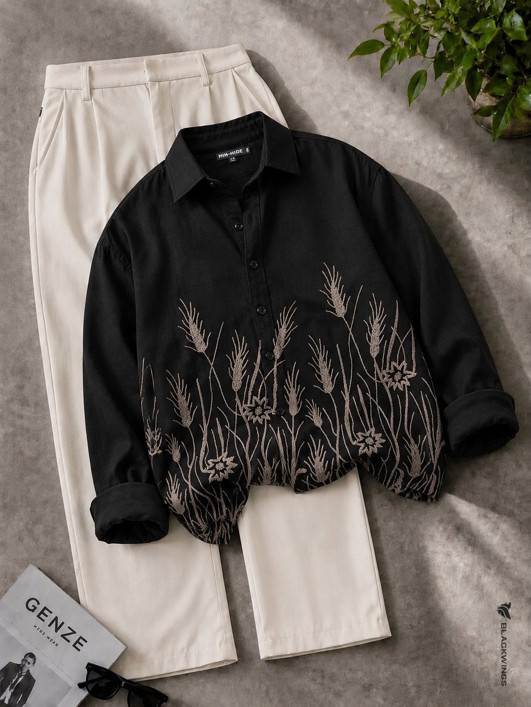 Black patterned shirt with beige floral designs laid over cream trousers on a grey background.