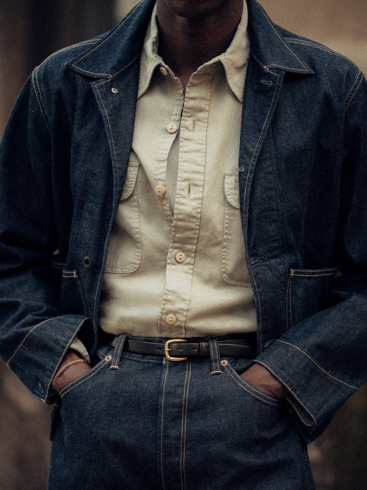 A person wearing a dark denim jacket, cream shirt, and blue denim jeans with a black belt.
