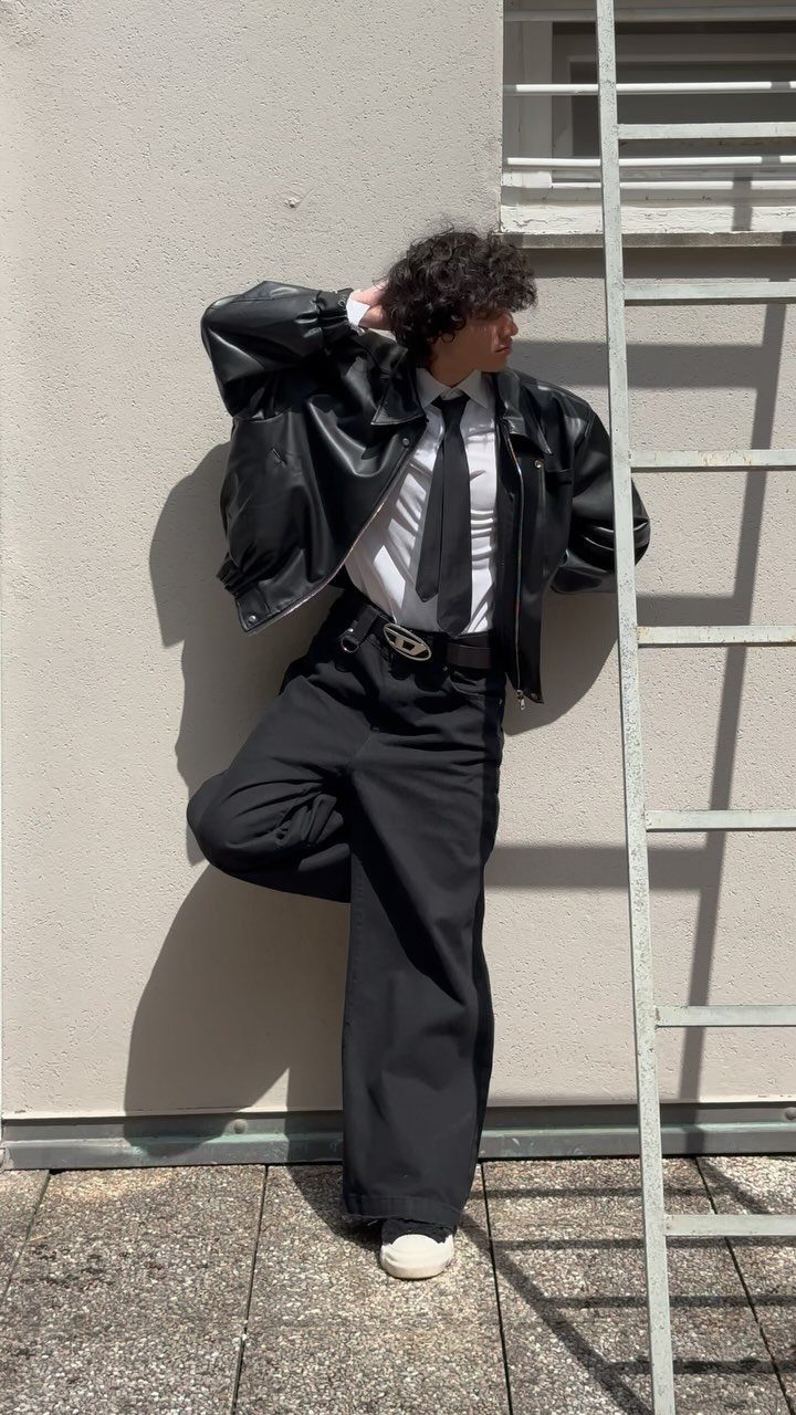 A person in a black leather jacket, white shirt, tie, and black trousers posing by a ladder.